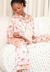 Rosa satinpyjamas med festliga blommiga och ornamentmönster, med krage och knapplås. Barnet håller en bok medan det sitter.