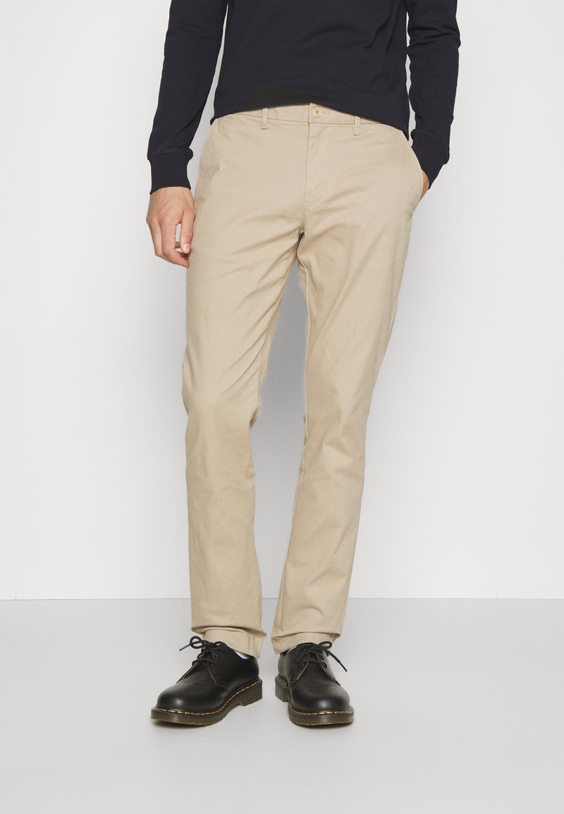 Pantaloni chinos color sabbia con taglio dritto, realizzati in tessuto di cotone liscio, abbinati a scarpe nere in pelle e a una maglia scura.