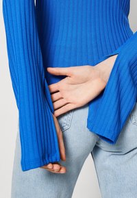Blue ribbed top with wide sleeves, featuring a fitted bodice. The texture is smooth; the sleeves have a slight flare, complementing the body.