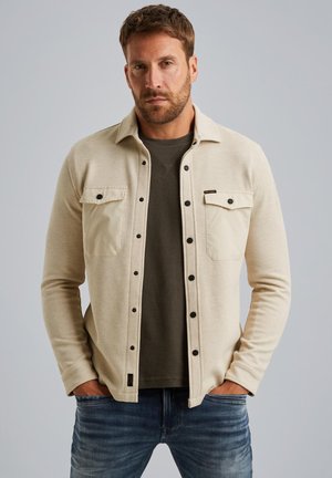 Beige strukturierte Shirtjacke mit zwei Fronttaschen, schwarzen Knöpfen und einem runden Kragen, über einem dunkelgrünen Sweatshirt getragen. Jeansstoff-Hose sichtbar.