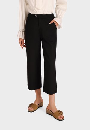 Pantaloni neri cropped con una texture liscia, dotati di chiusura frontale con bottone e tasche laterali, abbinati a sandali marroni slip-on con un accento floreale.