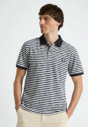 Mężczyzna w czarno-białej, poziomo paskowanej koszulce polo z czarnym kołnierzykiem i beżowych spodniach, stoi z rękami w kieszeniach.