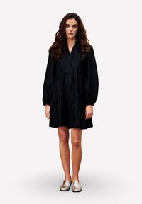Femme aux longs cheveux bouclés portant une robe noire à manches longues ample et des mocassins argentés, se tenant devant un fond blanc uni.
