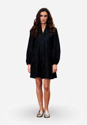 Femme aux longs cheveux bouclés portant une robe noire à manches longues ample et des mocassins argentés, se tenant devant un fond blanc uni.