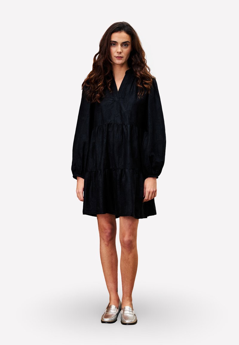Femme aux longs cheveux bouclés portant une robe noire à manches longues ample et des mocassins argentés, se tenant devant un fond blanc uni.
