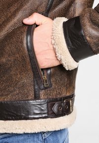 Veste en cuir marron avec une finition texturée, col en peau lainée, poche zippée et détail de taille ceinturée.