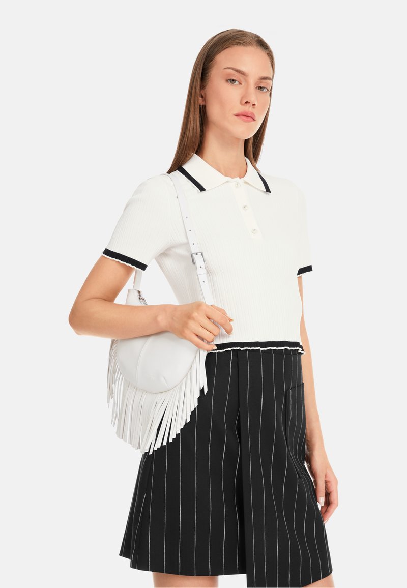 Weißes gestreiftes Polohemd mit schwarzem Kragen und Akzenten, kombiniert mit gestreiften schwarzen Shorts. Das Model hält eine weiße Fransen-Handtasche.