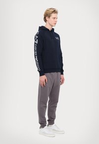 Kék kapucnis pulóver, fehér logónyomatokkal az ujjakon, szürke melegítőnadrággal párosítva. A modell fehér sportcipőt visel, világos háttér előtt áll.