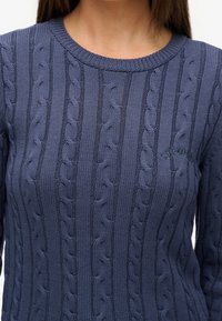 Pull en tricot bleu marine avec texture torsadée, col rond côtelé et logo brodé discret sur la poitrine. Manches longues.