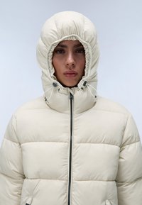Beige pufferjack met een hoge kraag en een trekkoordkap. Voorzien van horizontale stiksels en een voormetaalrits. Gladde, vlakke stof.