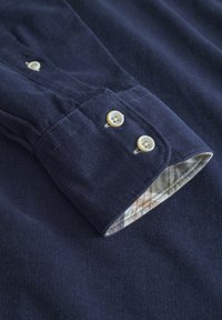 Polsino della camicia in corduroy blu navy con due bottoni chiari. L'interno del polsino presenta un motivo a quadretti nelle tonalità beige, blu e arancione.