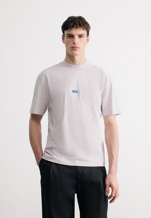 Νεαρός με κοντά σκούρα μαλλιά που φοράει ένα ανοιχτό μπεζ κοντομάνικο T-shirt με μικρό μπλε λογότυπο "Hugo" και μαύρο παντελόνι.