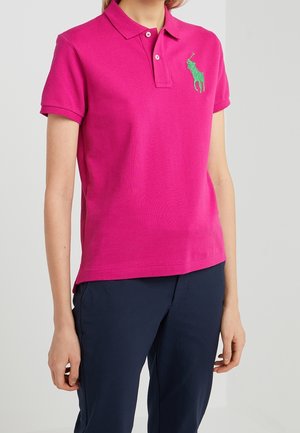 Kobieta w jasnoróżowej polo z krótkim rękawem z zielonym logo na piersi i ciemnogranatowych spodniach, stojąca na jednolitym tle.