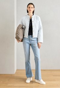 Chaqueta acolchada de color azul claro con botones dorados, camiseta negra, jeans de corte acampanado de lavado claro y zapatillas blancas. Bolsa gris sostenida en la mano izquierda.