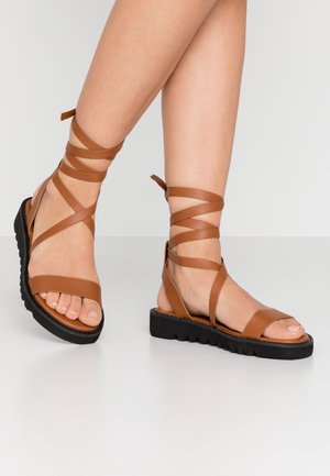 Sandales en cuir marron avec des lanières entrecroisées à la cheville, design à bout ouvert et une semelle en caoutchouc noir texturé.