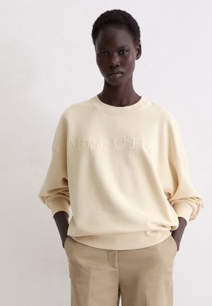 Beige oversized sweatshirt with stitched logo, round neckline, and dropped shoulders. Paired with beige pants, smooth texture.