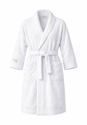 Frau mit langen blonden Haaren, die einen weißen Plüschbademantel mit Taschen und einem gebundenen Gürtel vor einem schlichten hellen Hintergrund trägt.