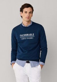 Homme portant un sweat-shirt bleu marine avec le texte "FAÇONNABLE CÔTE D'AZUR", une chemise rayée bleue et blanche en dessous, et un pantalon blanc, les mains dans les poches.