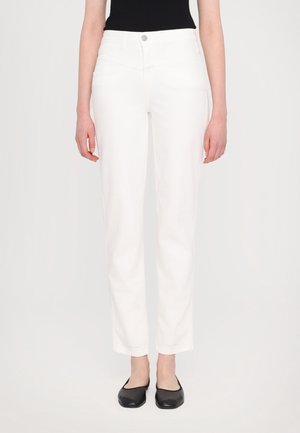 Kobieta w prostych białych spodniach i czarnych balerinach stoi na tle jednolitego, jasnego tła.