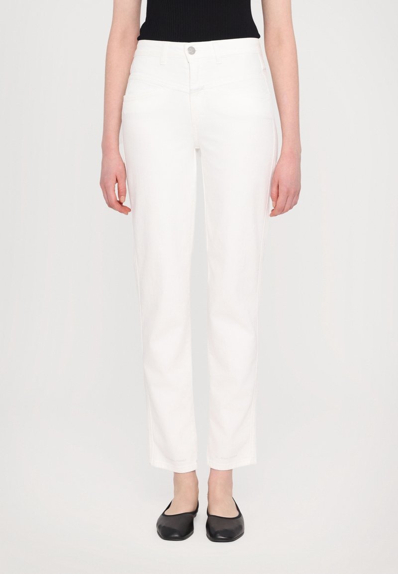 Femme portant un pantalon blanc droit et des ballerines noires, debout devant un fond uni clair.