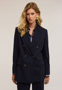 Granatowa marynarka dwurzędowa z złotymi guzikami, dwiema przednimi kieszeniami i wyraźnie zarysowanymi ramionami. Założona na wielokolorową koszulę z kołnierzykiem.