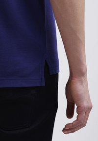 Bras et main d'une personne portant une chemise bleue et un pantalon noir, vus de profil sur un fond uni.