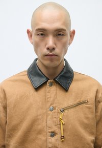 Brun kanvasjakke med svart lærkrage, med en glidelås brystlomme og et gult bånd som aksent, designet med metallknapper.