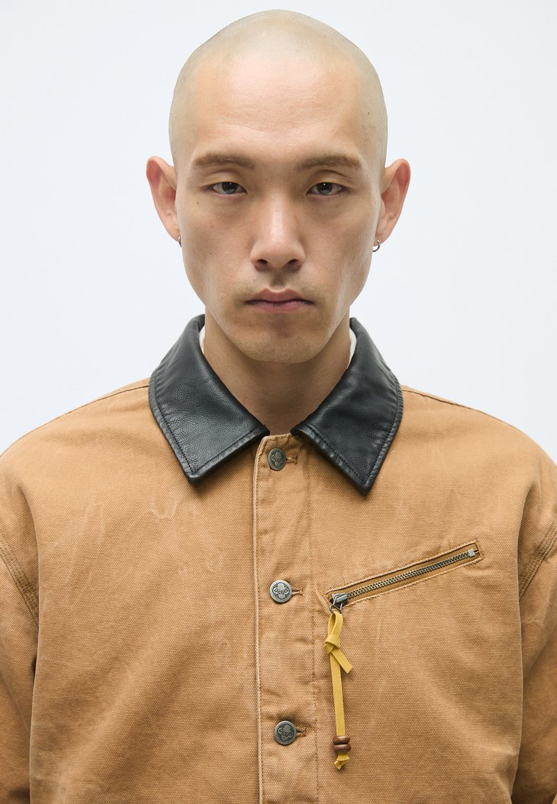 Braune Canvas-Jacke mit einem schwarzen Lederkragen, versehen mit einer mit Reißverschluss versehenen Brusttasche und einem gelben Band, gestaltet mit Metallknöpfen.