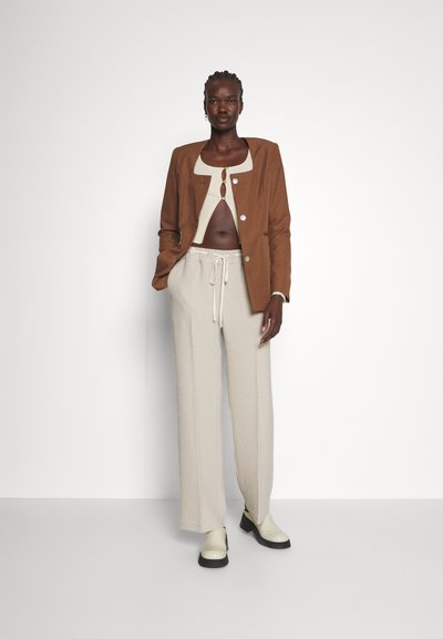 Γυναίκα με σκούρο δέρμα και πλεξούδες φοράει καφέ σακάκι, κρεμ κοντό ζακέτα, μπεζ παντελόνι με κορδόνια και κρεμ μποτάκια αστραγάλου σε λευκό φόντο.