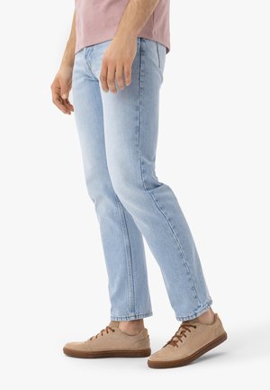 Mies pukeutuneena vaaleansinisiiin farkkuihin ja beigen värisiin rentoihin tennareihin, seisoo yksinkertaisen valkoisen taustan edessä.