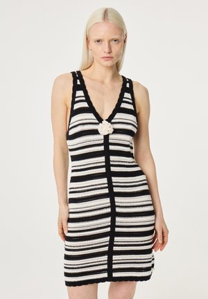 Vrouw draagt een zwart-wit gestreepte mouwloze gebreide jurk met een diepe V-hals en een wit bloemdetail in het midden.