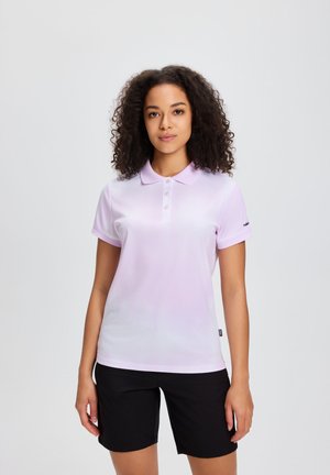 Vrouw met krullend haar, gekleed in een lichtpaarse polo met korte mouwen en zwarte korte broek tot op de knie, staand tegen een effen achtergrond.