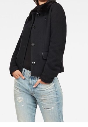 Femme portant une veste noire boutonnée avec un col en fourrure et un jean bleu clair déchiré, mains dans les poches, sur un fond blanc.