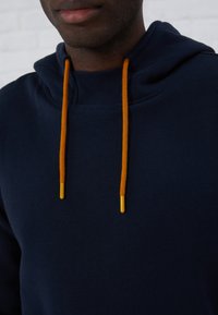 Sudadera azul marino de tejido texturizado, con cordones naranjas y puntas amarillas. Diseño simple, cuello redondeado y capucha ajustable.