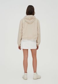 Chaqueta beige con capucha, con puños y dobladillo elásticos, combinada con una falda de mezclilla blanca. Las botas son de color claro, complementando el conjunto.