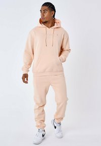 Perzikkleurige hoodie met kangoeroezak en trekkoord, gecombineerd met bijpassende joggers. Bevat geborduurde "PROJECT X" branding. Witte sneakers.