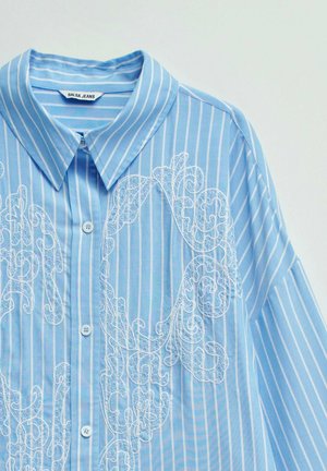 Chemise bleu clair à manches longues avec des rayures verticales blanches et des volutes brodées blanches complexes, fermeture à boutons sur le devant et col pointu.