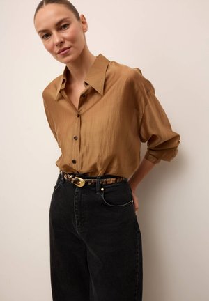 Mujer con el pelo recogido llevando una camisa marrón holgada abotonada metida en unos vaqueros negros de cintura alta con un cinturón estampado.