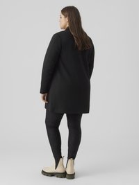 Μαύρο παλτό με δομημένο σχεδιασμό, σετ με μαύρο κολάν με υφή και κυματοειδές μοτίβο, και εκρού μποτάκια αστραγάλου με πράσινες σόλες.