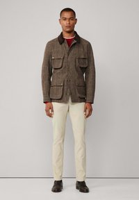 Bruine houndstooth jas met vier voorkruiszakken, een bruine kraag en knoopsluiting, gecombineerd met een lichte broek en donkere schoenen.