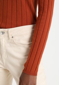 Haut côtelé à manches longues couleur rouille avec une coupe ajustée, associé à un jean taille haute couleur crème, doté d'une fermeture à boutons et de poches latérales.