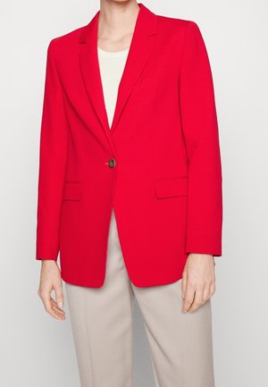 Blazer texturé rouge avec une fermeture à un bouton, revers cranté et poches avant. Porté par-dessus un haut clair et associé à un pantalon beige.
