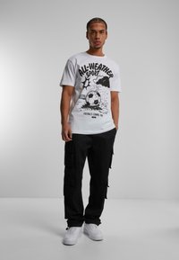 Hvid grafisk t-shirt med teksten "ALL-WEATHER SPORT" og fodbolddesign, kombineret med sorte cargo-bukser og hvide sneakers.