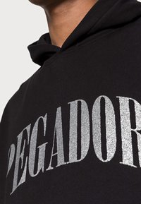 Felpa nera con stampa in glitter argento che mostra la parola "PEGADOR." Caratterizzata da un cappuccio con cordino e una texture di tessuto morbido.