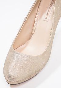 Escarpin à talons hauts doré pailleté avec un bout rond, doublure intérieure lisse et surface métallique texturée, offrant une silhouette élégante.