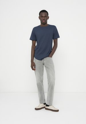 Granatowy, krótki rękaw t-shirt w połączeniu z jasnoszarymi, prostymi dżinsami oraz beżowymi tenisówkami, na tle jednolitego białego tła.