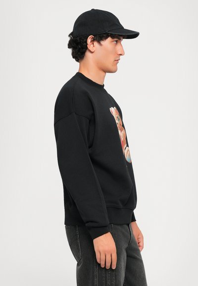 Giovane uomo con un cappellino nero, felpa nera con un grafica di un orsetto e jeans scuri, in piedi di fronte a destra su uno sfondo bianco uniforme.