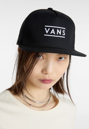 Gorra negra de Vans con visera plana; presenta un logotipo bordado en blanco. Tejido suave con un diseño estructurado, llevada con una camiseta de color claro.