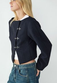 Veste courte navy en tissu texturé, avec des manches longues et des fermetures en métal argenté à l'avant. Associée à un jean bleu clair.