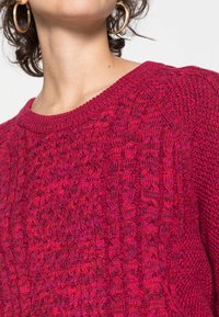 Sweter dzianinowy w intensywnym różowym odcieniu, z fakturowanym wzorem i ściągaczem przy dekolcie. Zbliżenie ukazuje szczegółową fakturę materiału.